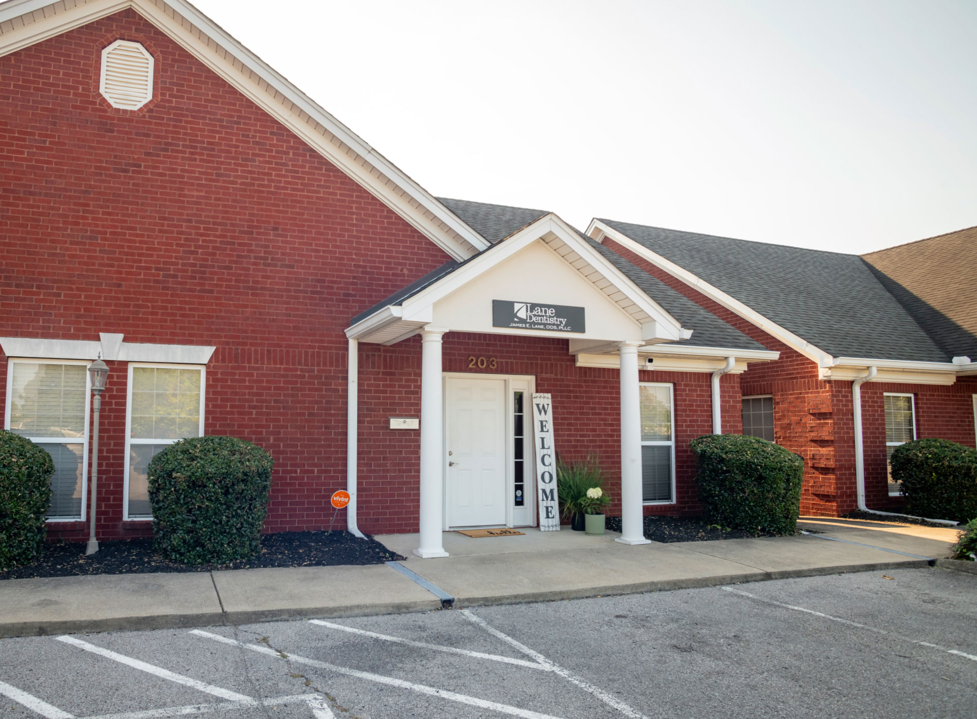Lane Dentistry red brick building exterior with white trim, small porch cover, welcome sign, and green hedges