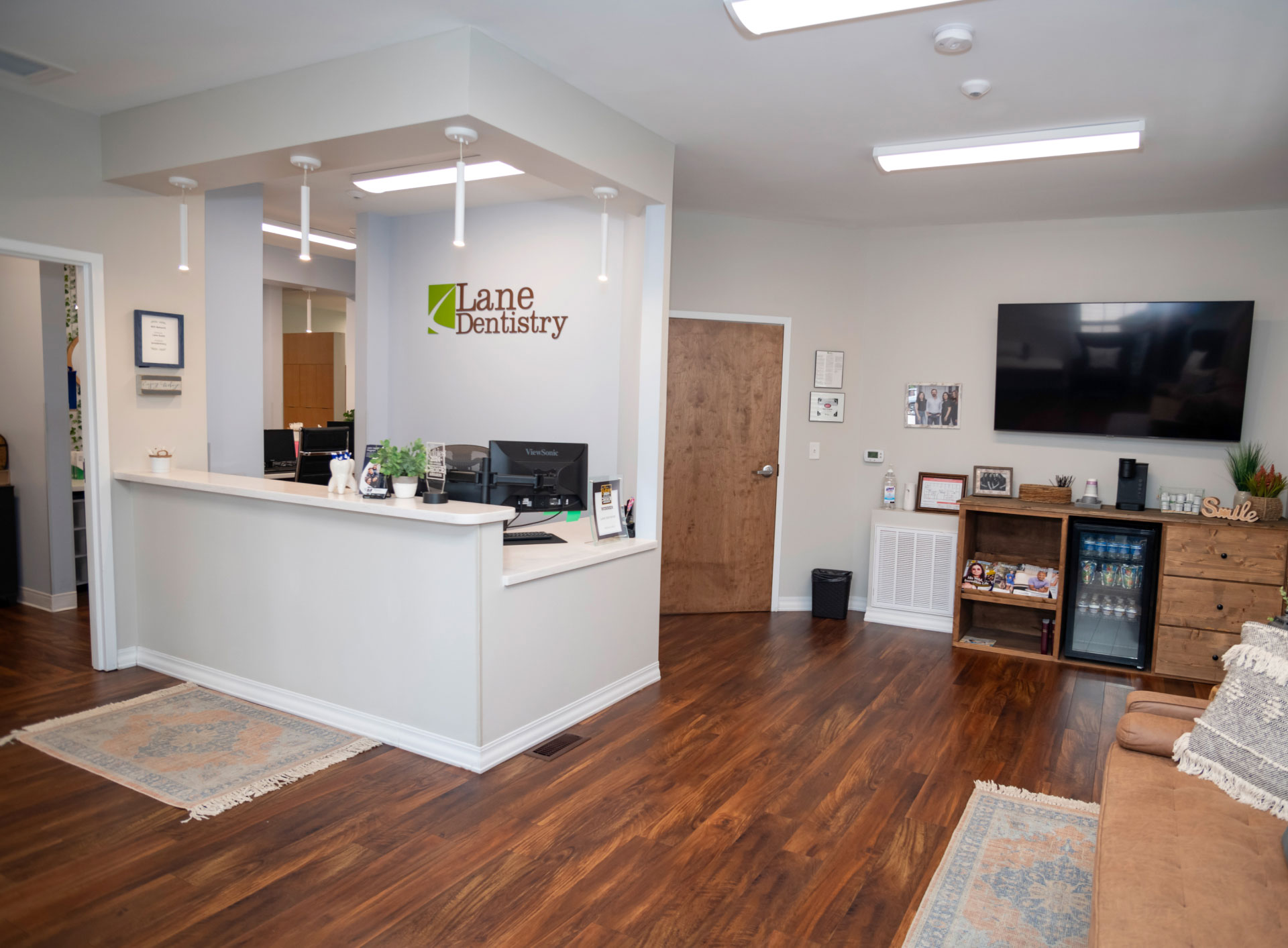 front desk area with Lane Dentistry logo on wall, brown wood floors, and TV hanging on wall over table with mini fridge and coffee pot
