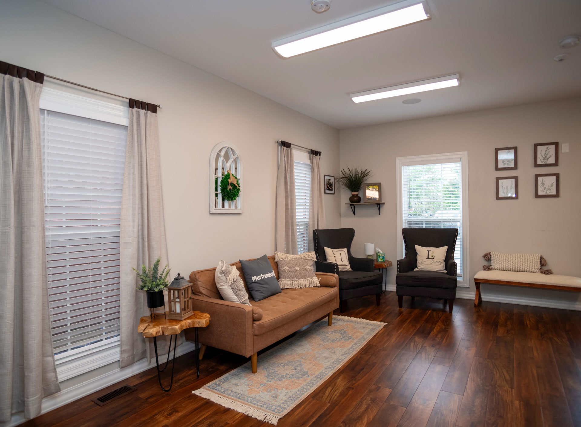 warm and inviting reception area with brown couch and gray wingback chairs for seating, 3 windows, and brown wood floors